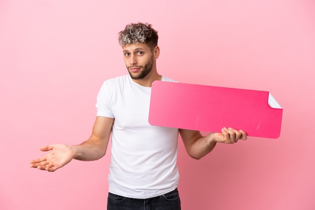 Giovane uomo caucasico bello isolato su sfondo rosa che tiene un cartello vuoto e ha dei dubbi