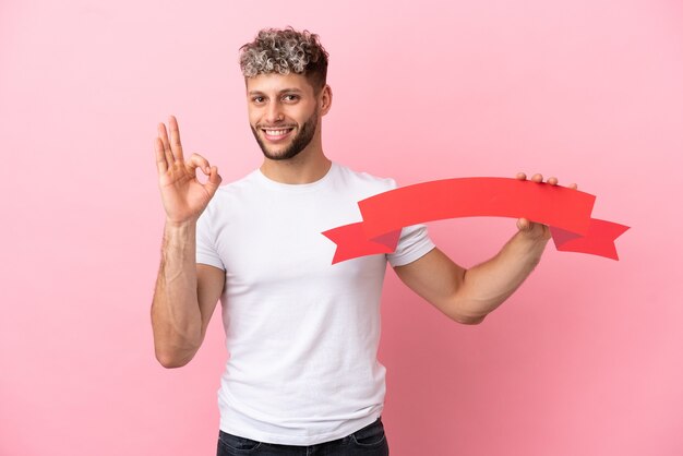 Giovane uomo caucasico bello isolato su sfondo rosa con in mano un cartello vuoto e facendo segno OK