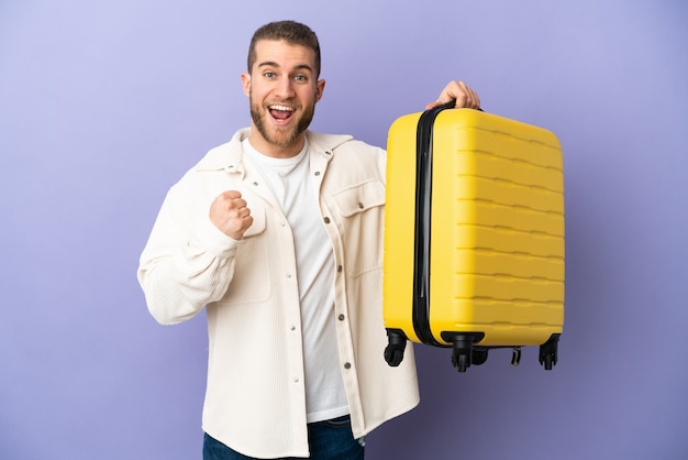Giovane uomo caucasico bello isolato su viola in vacanza con la valigia di viaggio