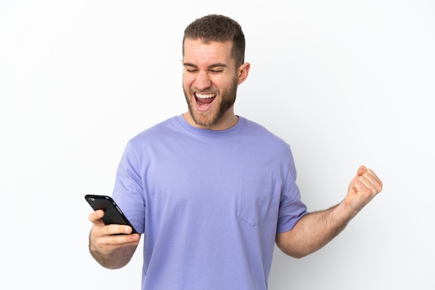 Giovane uomo caucasico bello isolato sul muro bianco utilizzando il telefono cellulare e facendo il gesto di vittoria