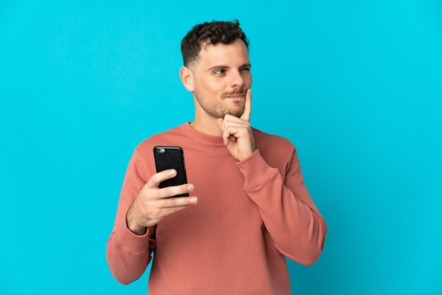 Giovane uomo caucasico bello isolato sull'azzurro utilizzando il telefono cellulare e il pensiero