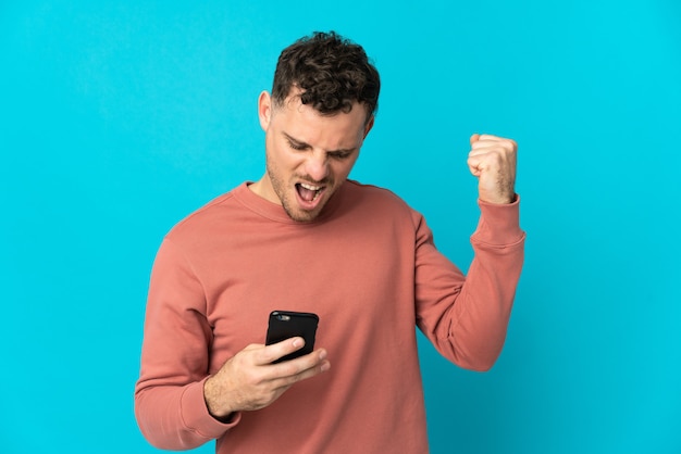 Giovane uomo caucasico bello isolato sulla parete blu utilizzando il telefono cellulare e facendo il gesto di vittoria