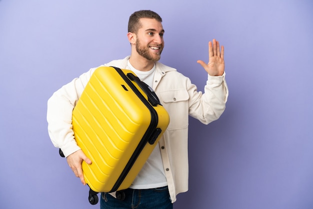 Giovane uomo caucasico bello isolato sulla parete viola in vacanza con la valigia di viaggio e salutando