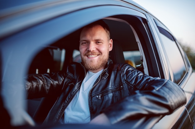 Giovane uomo caucasico biondo barbuto sorridente bello in giacca di pelle in posa nella sua automobile moderna.