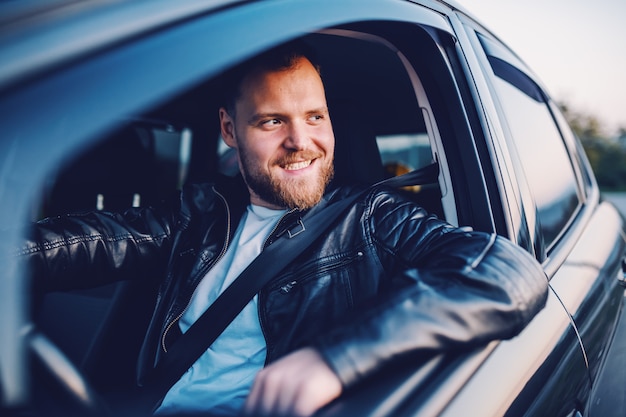 Giovane uomo caucasico biondo barbuto sorridente bello in giacca di pelle in posa nella sua automobile moderna.