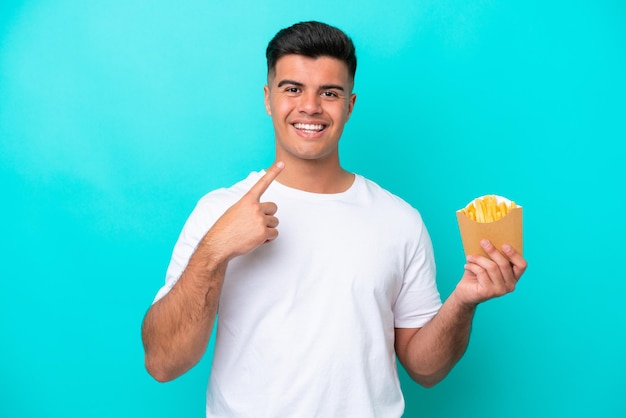 Giovane uomo caucasico che cattura patatine fritte isolate su sfondo blu dando un pollice in alto gesto