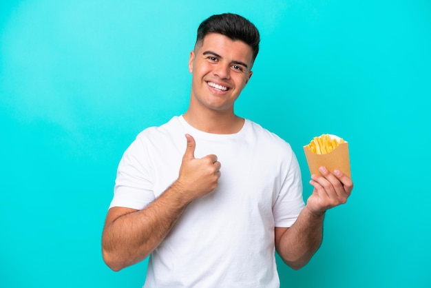 Giovane uomo caucasico che cattura patatine fritte isolate su sfondo blu dando un pollice in alto gesto