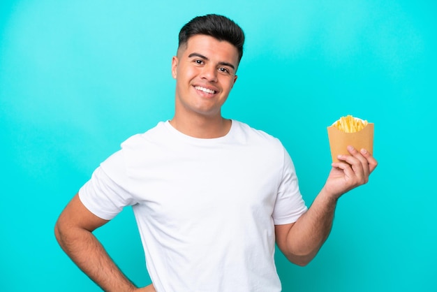 Giovane uomo caucasico che cattura patatine fritte isolate su sfondo blu in posa con le braccia all'anca e sorridente