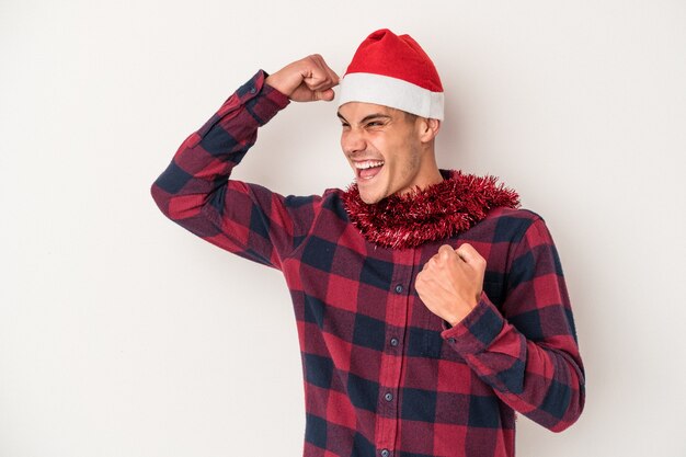 Giovane uomo caucasico che celebra il Natale isolato su sfondo bianco alzando il pugno dopo una vittoria, concetto vincitore.