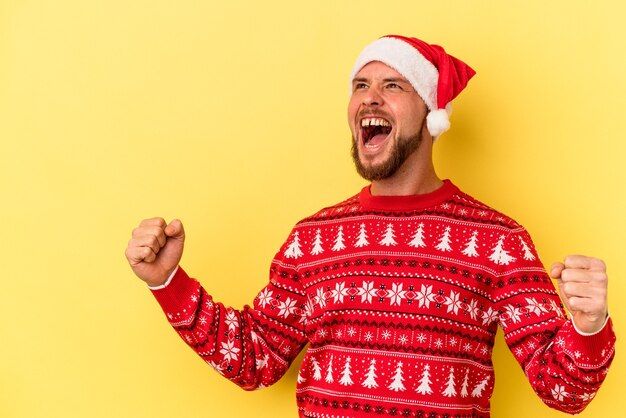 Giovane uomo caucasico che celebra il Natale isolato su sfondo giallo alzando il pugno dopo una vittoria, concetto vincitore.