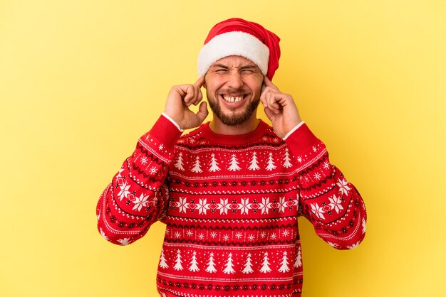 Giovane uomo caucasico che celebra il Natale isolato su sfondo giallo che copre le orecchie con le mani.