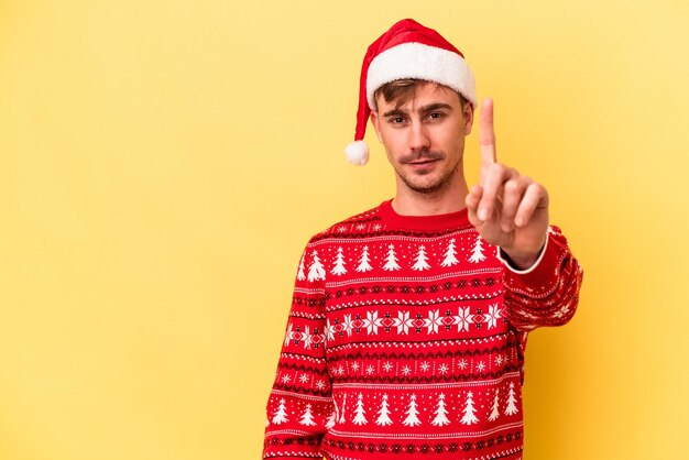 Giovane uomo caucasico che celebra il Natale isolato su sfondo giallo che mostra il numero uno con il dito.