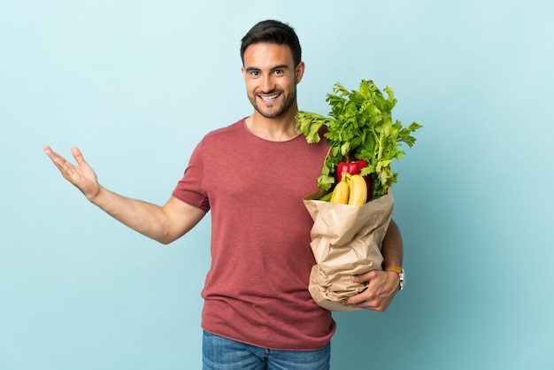 Giovane uomo caucasico che compra alcune verdure isolate sulla parete blu che estende le mani a lato per invitare a venire