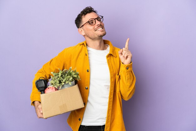 Giovane uomo caucasico che fa una mossa mentre prende in mano una scatola piena di cose isolate su viola che indica una grande idea