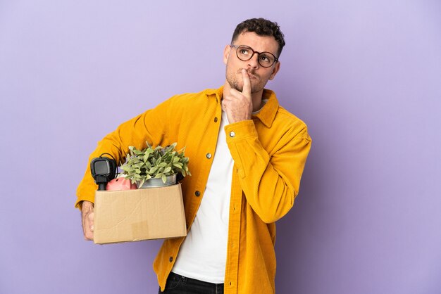 Giovane uomo caucasico che fa una mossa mentre si prende una scatola piena di cose isolate su sfondo viola che ha dubbi mentre si guarda in alto