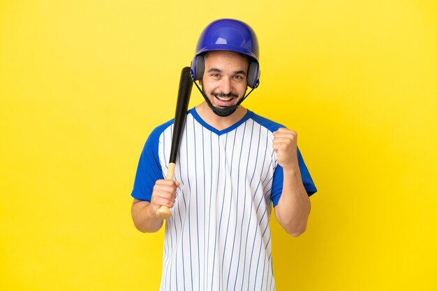 Giovane uomo caucasico che gioca a baseball isolato su sfondo giallo che celebra una vittoria nella posizione del vincitore