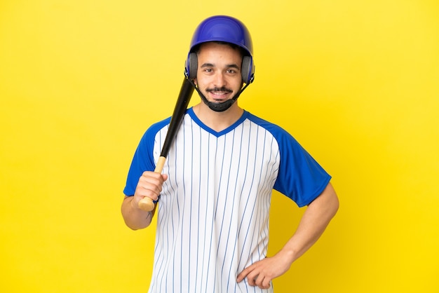 Giovane uomo caucasico che gioca a baseball isolato su sfondo giallo in posa con le braccia all'anca e sorridente