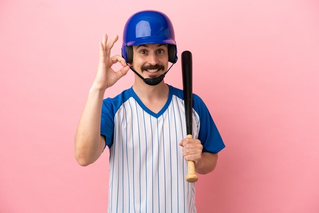 Giovane uomo caucasico che gioca a baseball isolato su sfondo rosa che mostra segno ok con le dita