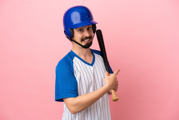 Giovane uomo caucasico che gioca a baseball isolato su sfondo rosa che punta indietro