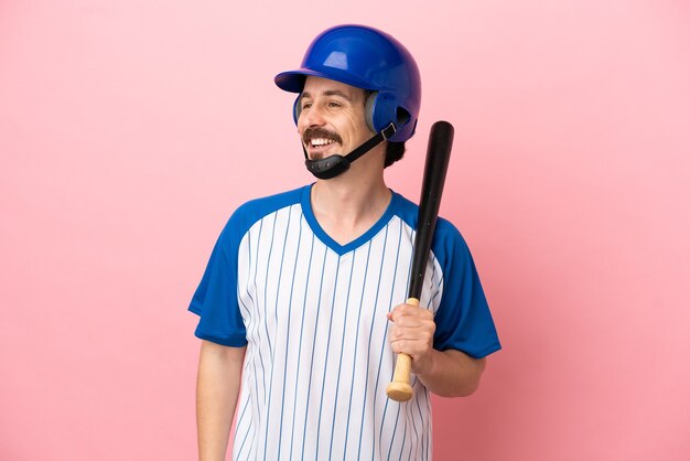 Giovane uomo caucasico che gioca a baseball isolato su sfondo rosa guardando di lato e sorridente