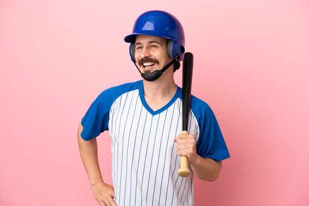 Giovane uomo caucasico che gioca a baseball isolato su sfondo rosa in posa con le braccia all'anca e sorridente