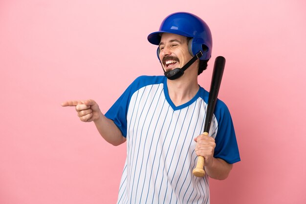 Giovane uomo caucasico che gioca a baseball isolato su sfondo rosa puntando il dito di lato e presentando un prodotto