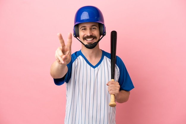 Giovane uomo caucasico che gioca a baseball isolato su sfondo rosa sorridendo e mostrando il segno della vittoria