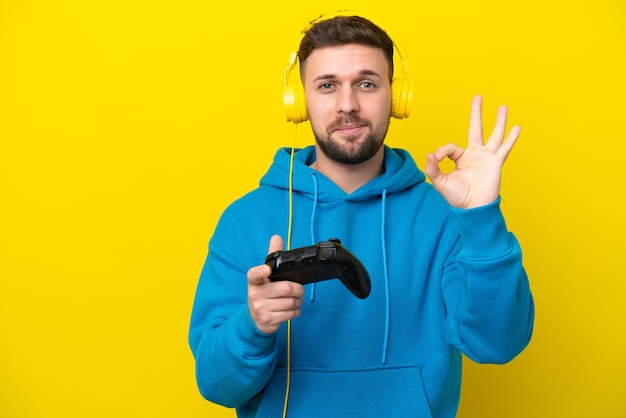 Giovane uomo caucasico che gioca con un controller per videogiochi isolato su sfondo giallo che mostra il segno giusto con le dita