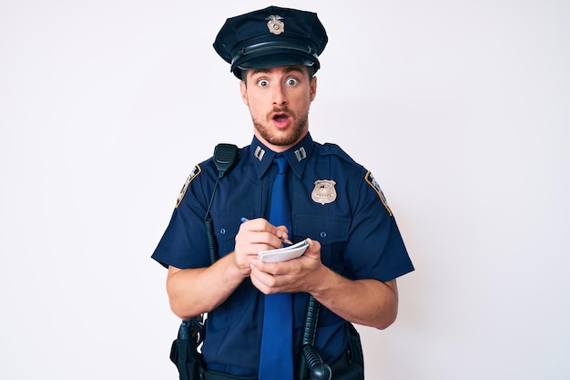 Giovane uomo caucasico che indossa l'uniforme della polizia che scrive bene in faccia d'urto che sembra scettico e sarcastico sorpreso con la bocca aperta