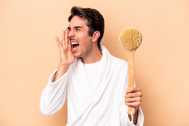 Giovane uomo caucasico che indossa un accappatoio che tiene un tiragraffi per la schiena isolato su sfondo beige che grida e tiene il palmo vicino alla bocca aperta.