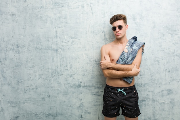 Giovane uomo caucasico che indossa un costume da bagno infelice guardando a porte chiuse con espressione sarcastica.