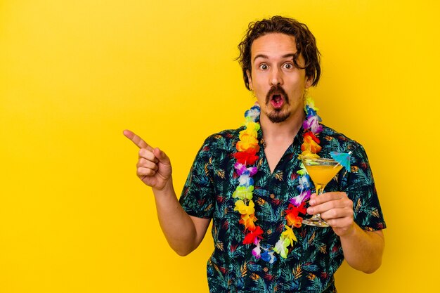 Giovane uomo caucasico che indossa una collana hawaiana che tiene un cocktail isolato sul muro giallo che punta di lato