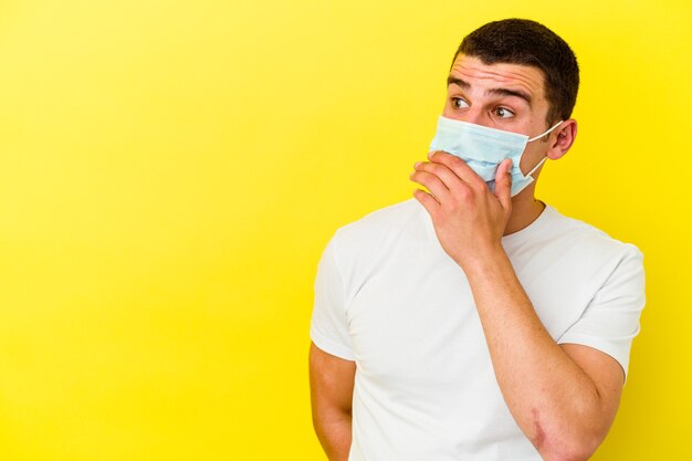 Giovane uomo caucasico che indossa una protezione per il coronavirus su giallo premuroso guardando uno spazio di copia che copre la bocca con la mano.