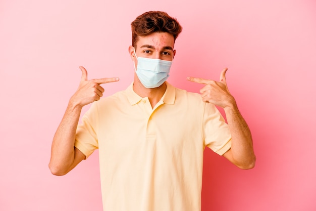 Giovane uomo caucasico che indossa una protezione per il coronavirus su sorrisi rosa, puntando le dita alla bocca.