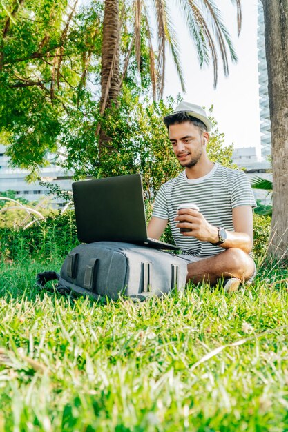 Giovane uomo caucasico che lavora con un laptop e tiene in mano un caffè seduto sul prato