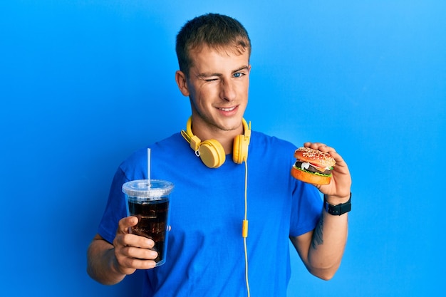 Giovane uomo caucasico che mangia un gustoso hamburger classico e una soda che strizza l'occhio guardando la telecamera con un'espressione sexy faccia allegra e felice