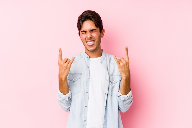 Giovane uomo caucasico che posa in una parete rosa che mostra gesto della roccia con le dita