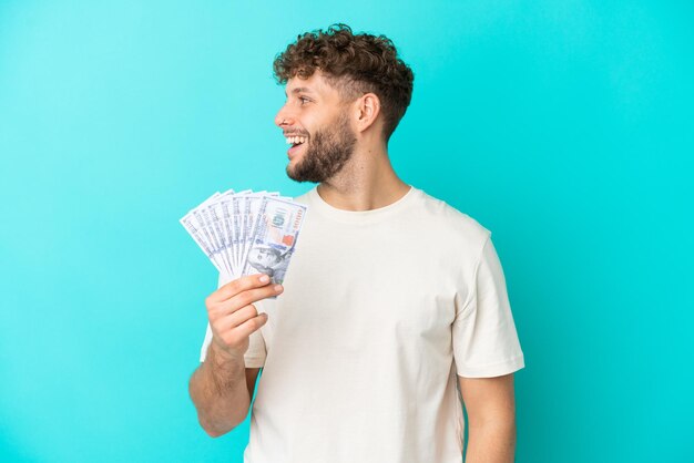 Giovane uomo caucasico che prende un sacco di soldi isolato su sfondo blu ridendo in posizione laterale