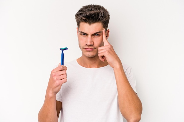 Giovane uomo caucasico che si rade la barba isolato su sfondo bianco che indica il tempio con il dito, pensando, concentrato su un compito.