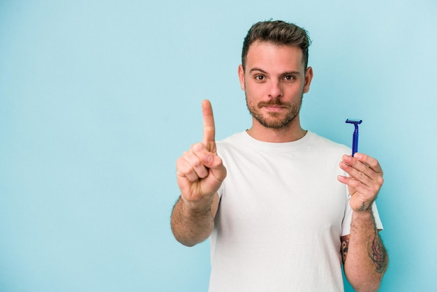 Giovane uomo caucasico che si rade la barba isolato su sfondo blu che mostra il numero uno con il dito.