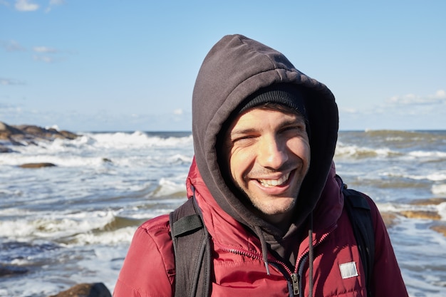 Giovane uomo caucasico che sorride alla macchina fotografica sulla spiaggia in inverno. Indossa giacca e cappuccio.