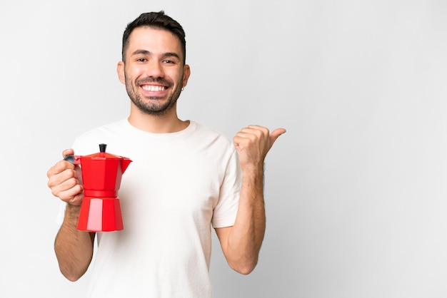 Giovane uomo caucasico che tiene caffettiera sopra fondo bianco isolato che indica il lato per presentare un prodotto