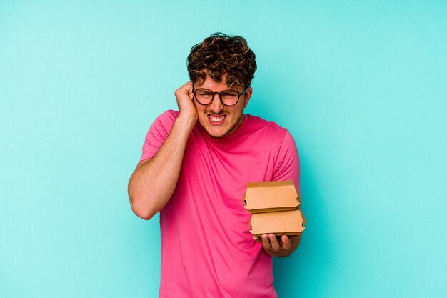 Giovane uomo caucasico che tiene due hamburger isolati su sfondo blu che copre le orecchie con le mani.