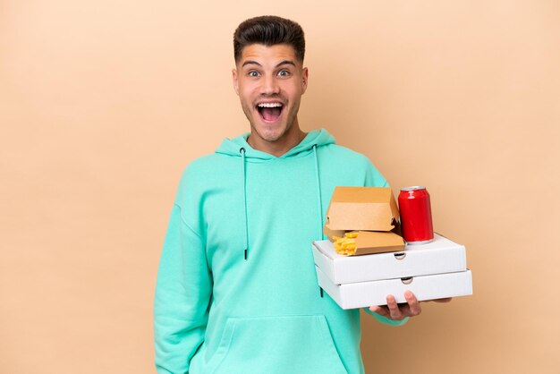 Giovane uomo caucasico che tiene fast food isolato su sfondo beige con espressione facciale a sorpresa