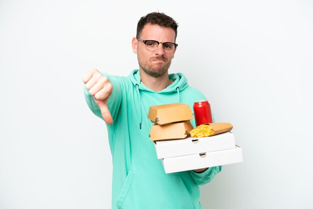 Giovane uomo caucasico che tiene fast food isolato su sfondo bianco che mostra il pollice verso il basso con espressione negativa