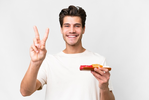 Giovane uomo caucasico che tiene il sashimi isolato su sfondo bianco sorridente e mostrando segno di vittoria