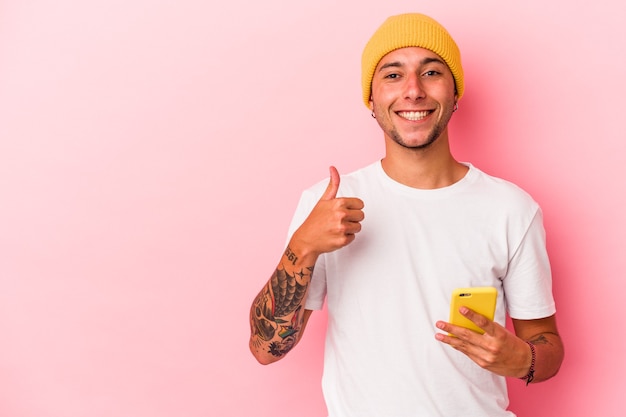 Giovane uomo caucasico che tiene il telefono cellulare isolato su sfondo rosa sorridente e alzando il pollice