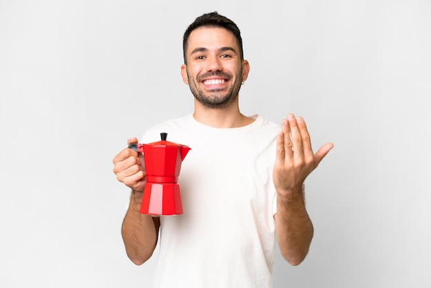 Giovane uomo caucasico che tiene la caffettiera su sfondo bianco isolato che invita a venire con la mano Felice che tu sia venuto