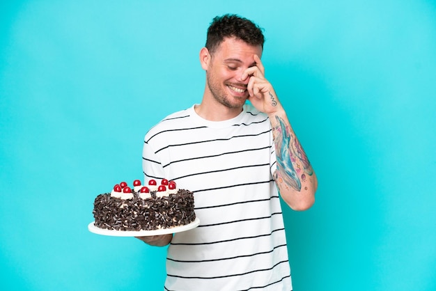 Giovane uomo caucasico che tiene la torta di compleanno isolata su sfondo blu ridendo