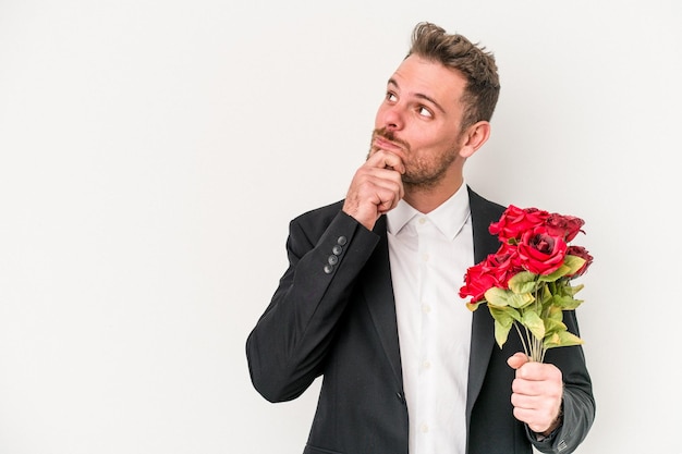 Giovane uomo caucasico che tiene mazzo di fiori isolato su sfondo bianco guardando lateralmente con espressione dubbiosa e scettica.